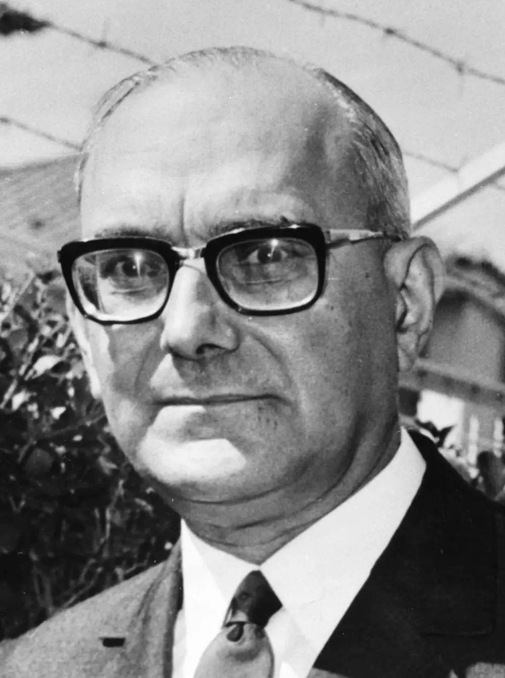 Black-and-white close-up portrait of a balding man in thick dark-framed glasses, white shirt, tie and dark jacket.