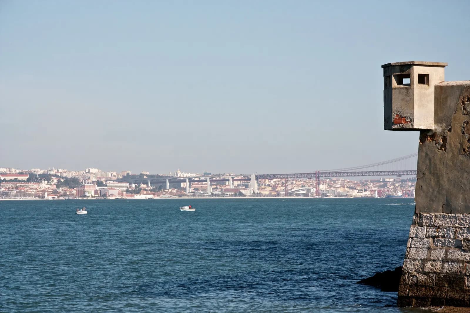 A weathered concrete sentry box perched on the corner of an old stone wall at the water's edge, with the wide blue river behind it and the Lisbon waterfront, a concrete viaduct and a long red suspension bridge on the far bank.