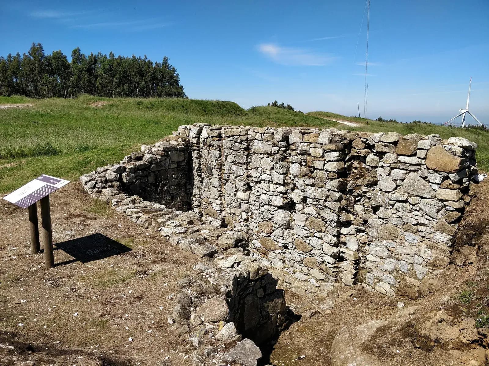 The excavated stone walls of a rectangular sunken room on a grassy hilltop, with an information panel on posts beside it and a wind turbine on the skyline.