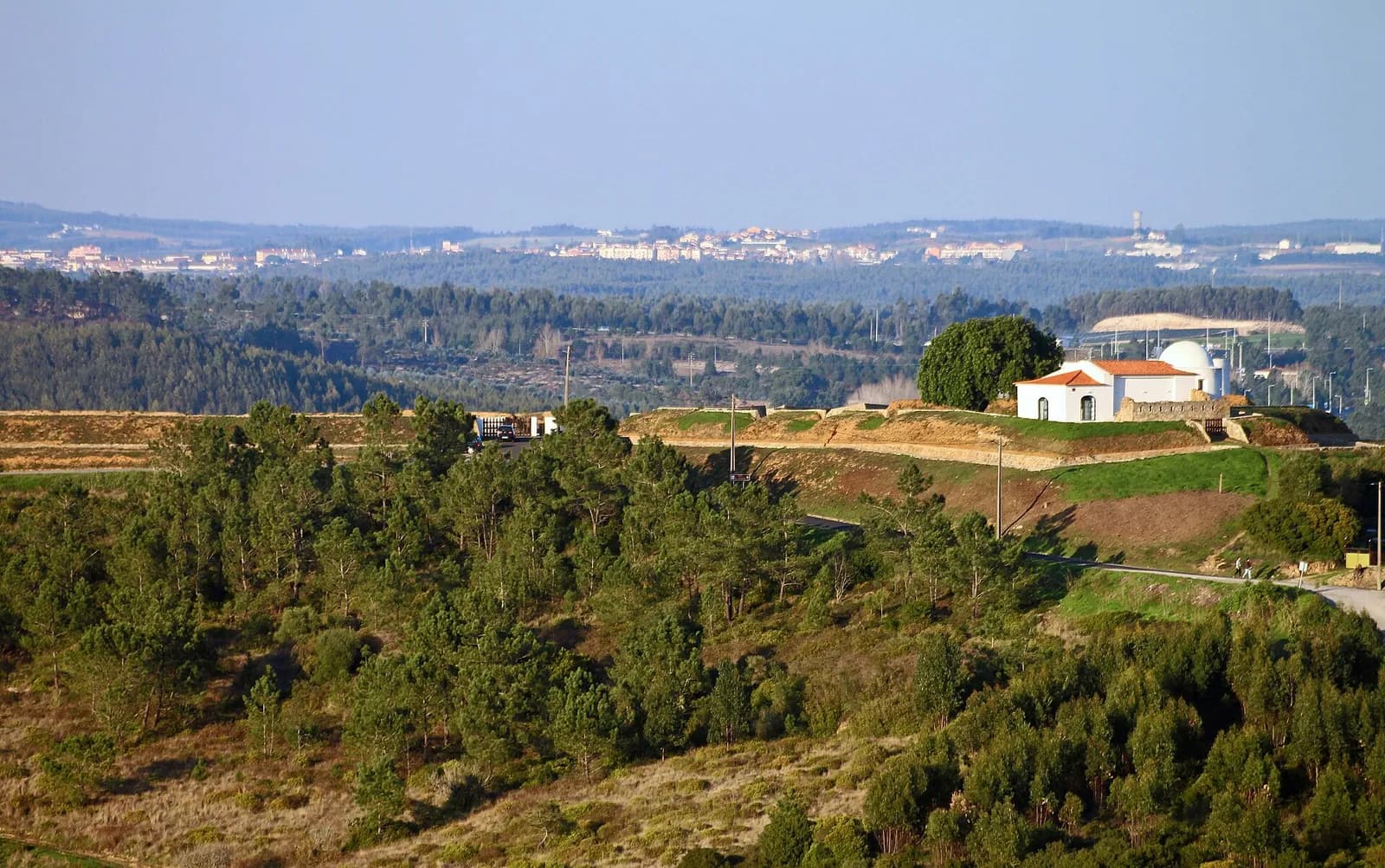 A hilltop covered in pine trees and grass, crowned by long low earth ramparts with a small white chapel and other white buildings on top; houses spread across the hills behind.