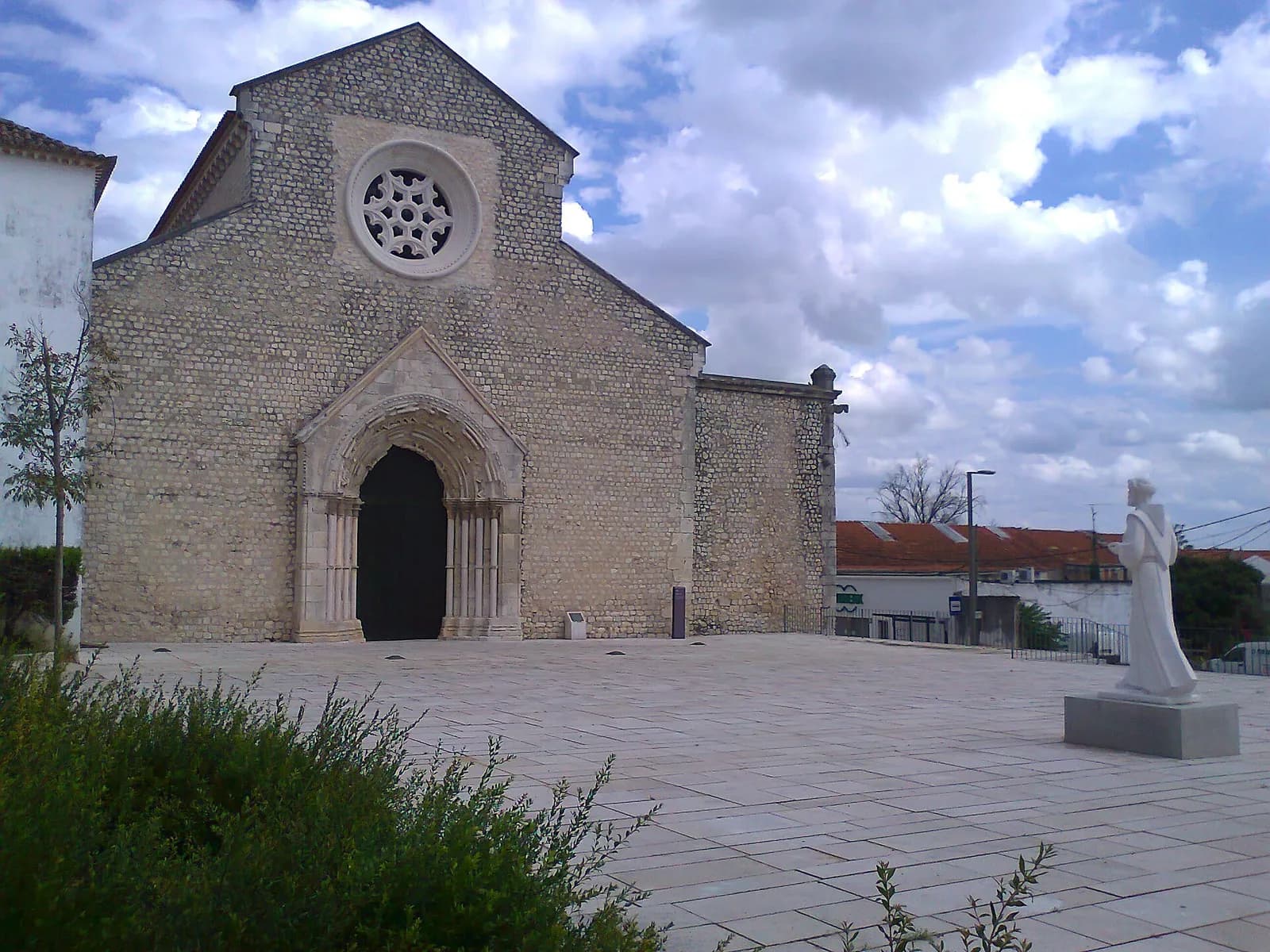 The plain stone front of a Gothic church with a round rose window above a deeply carved pointed doorway, facing a paved square with a white statue of a friar.