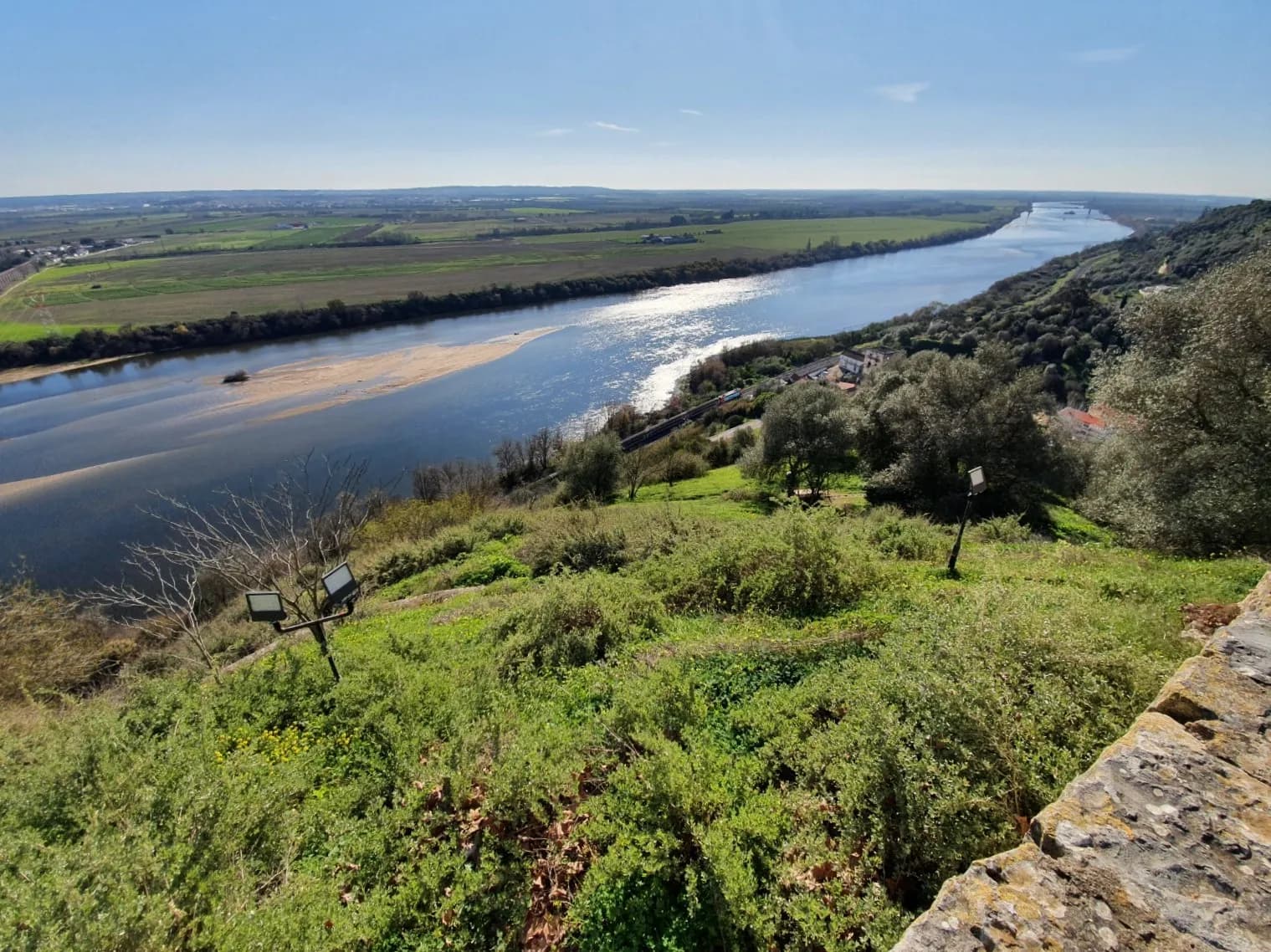 A wide river curves through a flat green plain of fields and lines of trees, seen from a high grassy slope with a stone wall in the foreground; a railway line and a few houses run along the near bank.