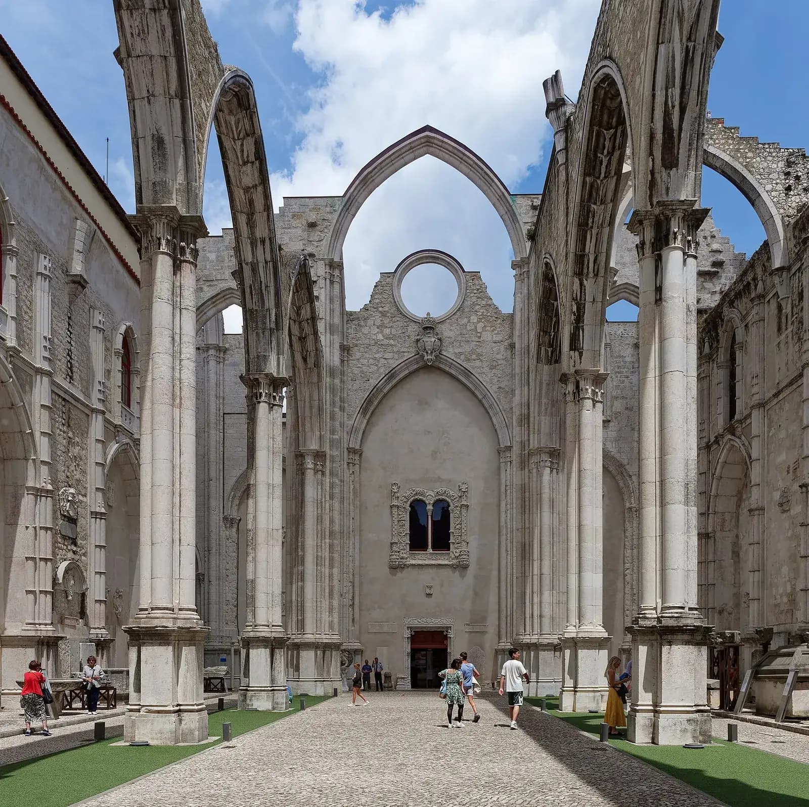 A roofless Gothic church nave seen from inside: tall pointed stone arches spring from slender pillars against a blue sky with clouds, over a paved central aisle with strips of grass and a few visitors.