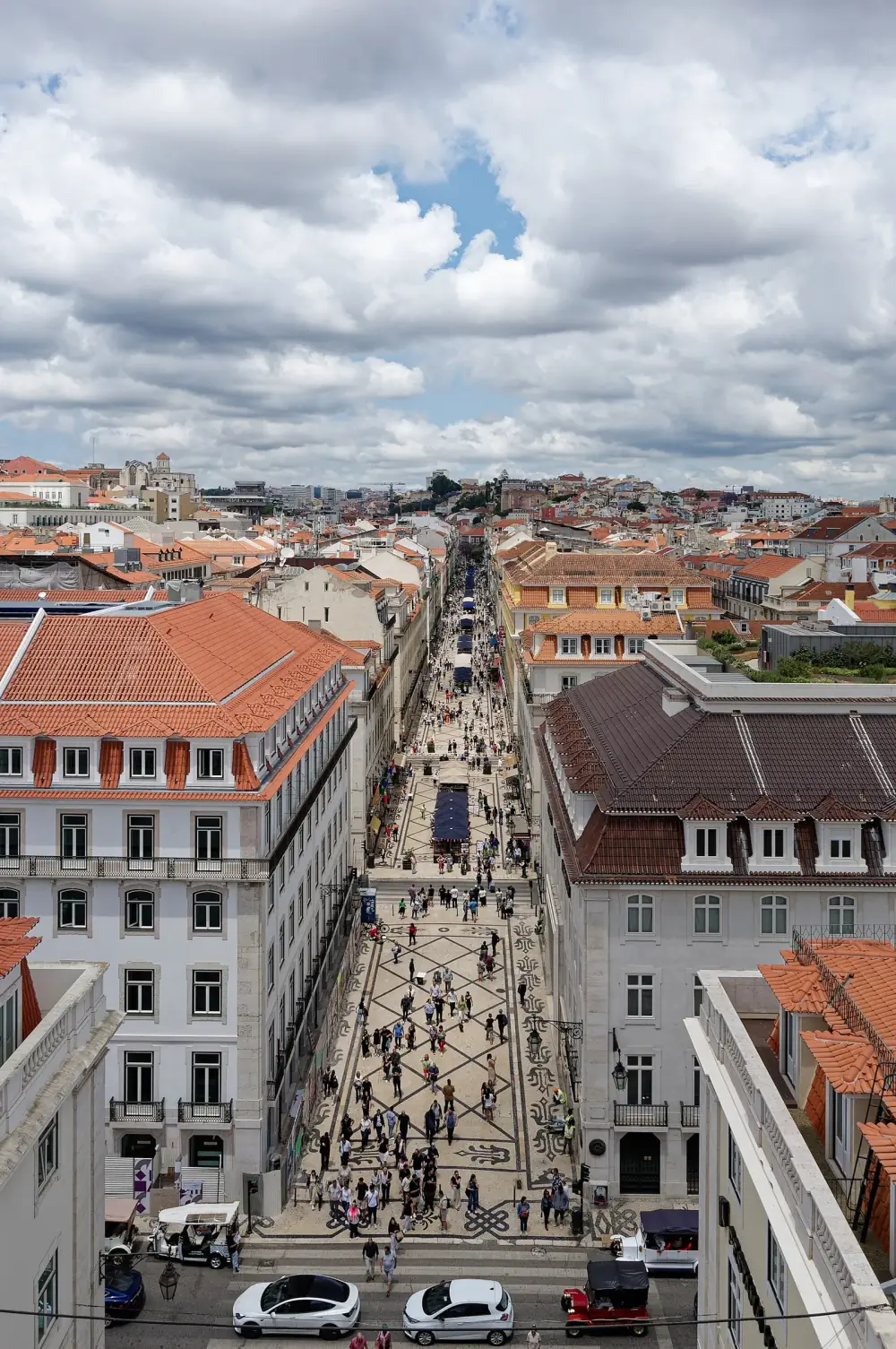View from a high arch down a long straight pedestrian street paved in patterned stone and busy with people, lined on both sides by uniform four- and five-storey buildings with regular rows of windows and red-tiled roofs.