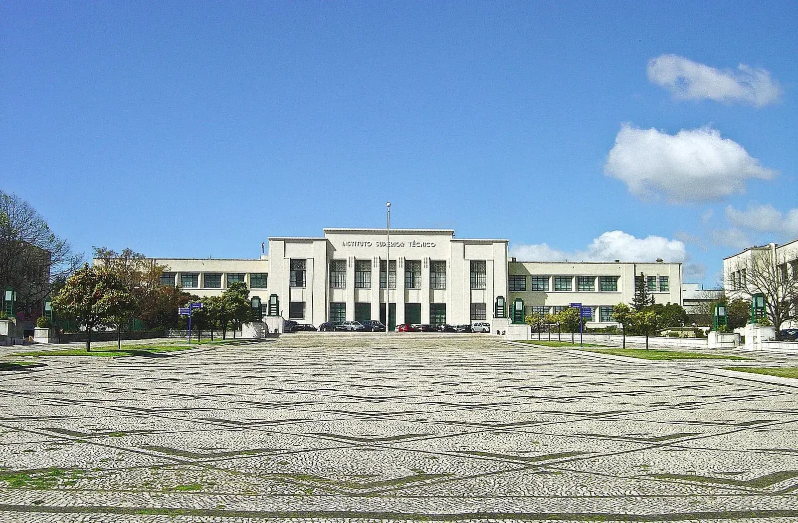 A long white modernist building with tall vertical windows and the words Instituto Superior Técnico over the entrance, seen across a wide square paved in black and white stone mosaic under a blue sky.