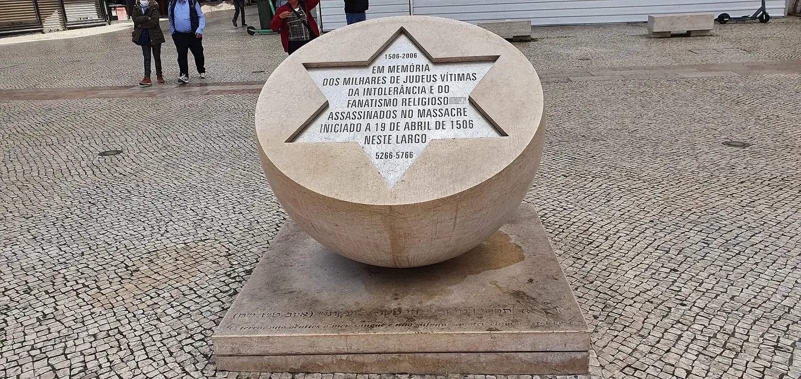 A pale stone hemisphere set on a square stone base on a cobbled square. Its flat, tilted top is cut with a six-pointed star filled with a Portuguese inscription, with the dates 1506–2006 above and 5266–5766 below.