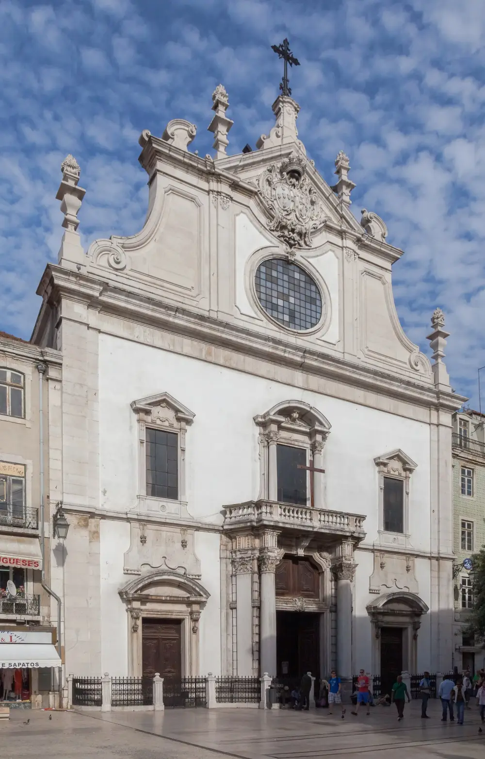 The white stone façade of a church under a blue sky, with a large oval window, a balcony over the central doorway, three doors behind a low railing and a few people on the square in front.