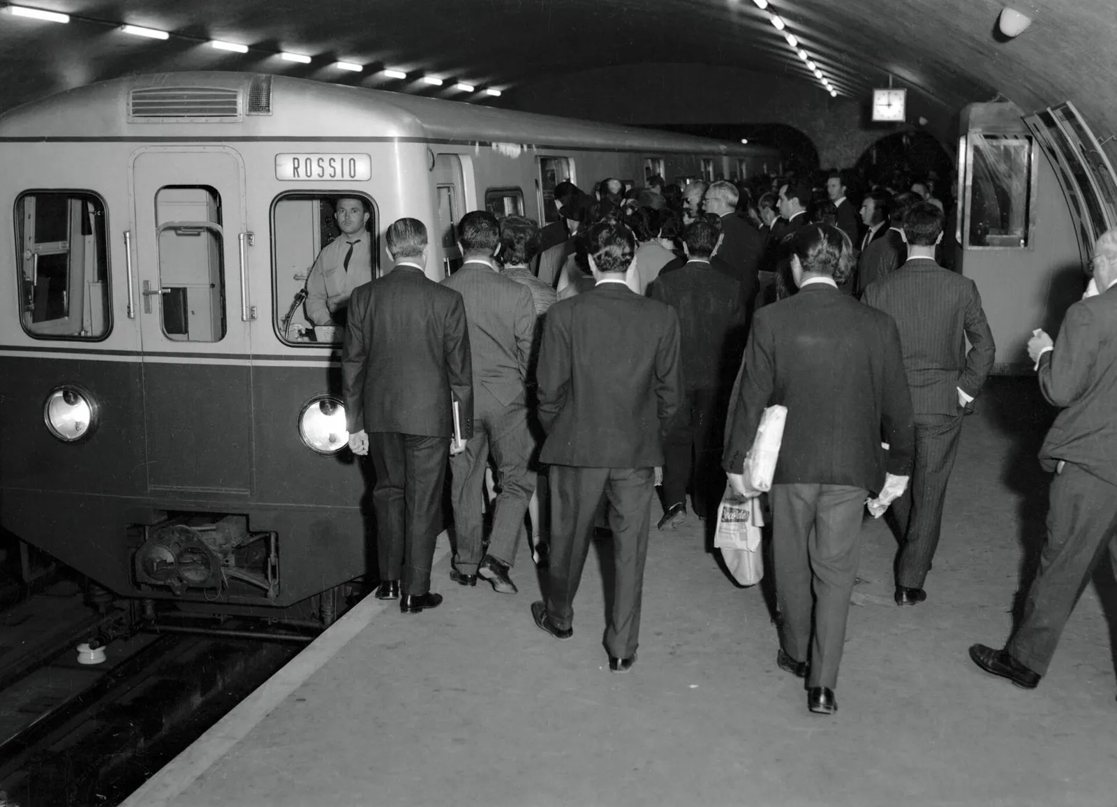 Black-and-white photograph of a crowd of men in dark suits, some carrying newspapers and parcels, walking along an underground platform beside a train whose destination board reads Rossio.