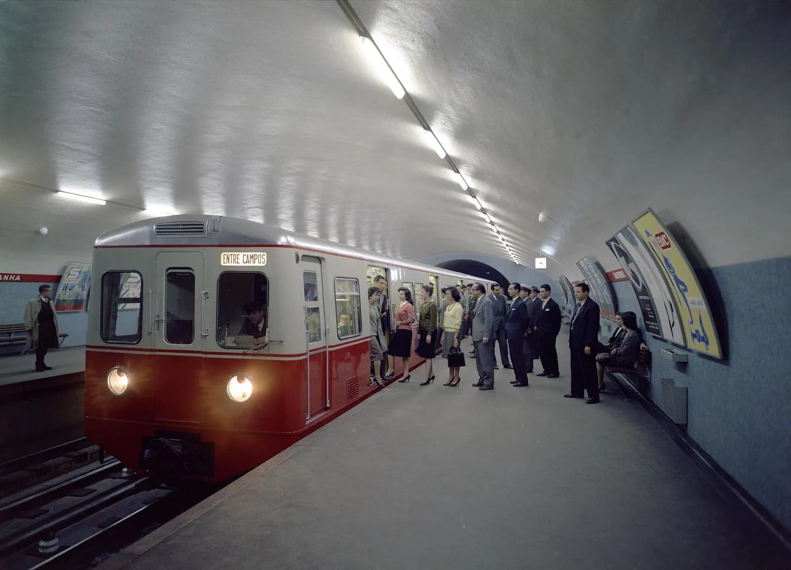 Colour photograph of a red-and-white train with a rounded front and a destination board reading Entre Campos at a curved, brightly lit underground platform. Men in suits and women in skirts step on and off through the open doors while the driver looks out of his cab.