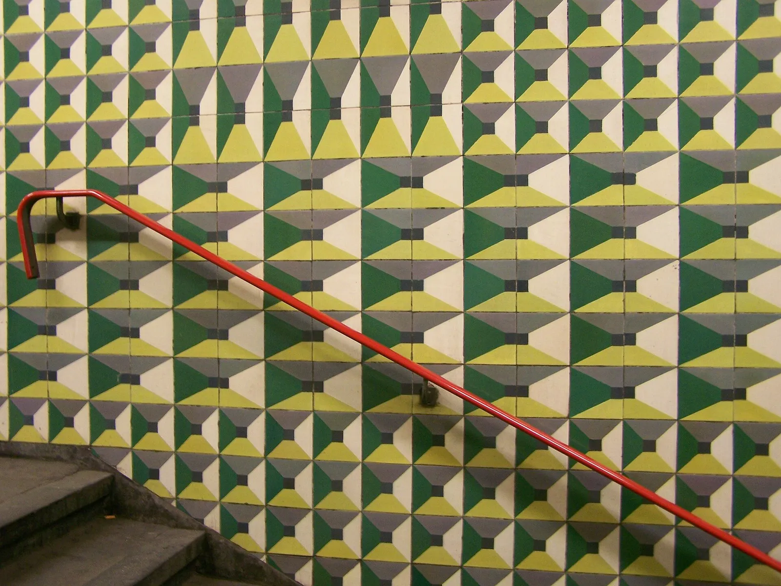 A staircase wall covered in square tiles forming rows of pyramid shapes in yellow, white, dark green and grey, with a red handrail running diagonally across it.