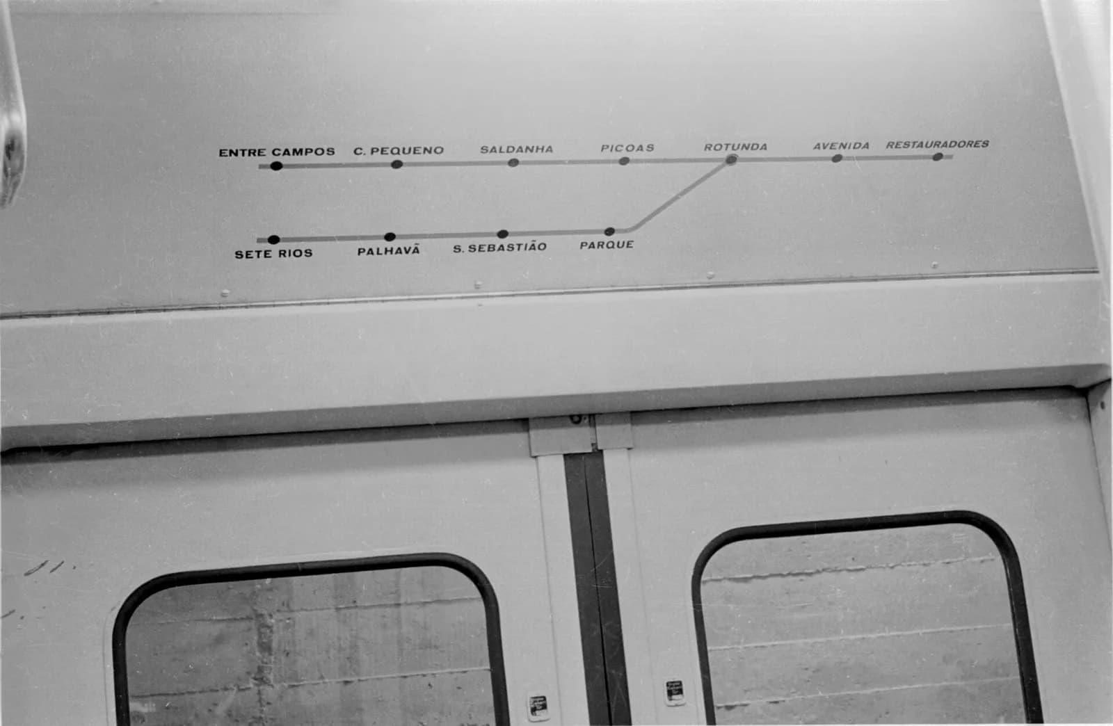 Black-and-white photograph of a line diagram painted above the sliding doors inside a carriage. An upper line runs from Entre Campos through C. Pequeno, Saldanha, Picoas and Rotunda to Avenida and Restauradores; a lower branch from Sete Rios through Palhavã, S. Sebastião and Parque joins it at Rotunda.