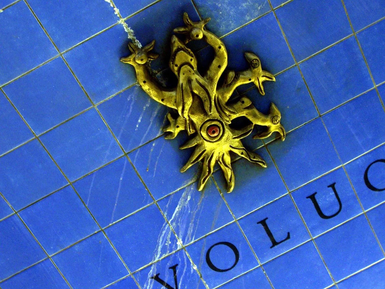 Close-up of a wall of deep blue square tiles with large black capital letters along the bottom and a gilded sculpture of a sun-like face with curling arms fixed to the tiles. White streaks of mineral deposit run down the wall.