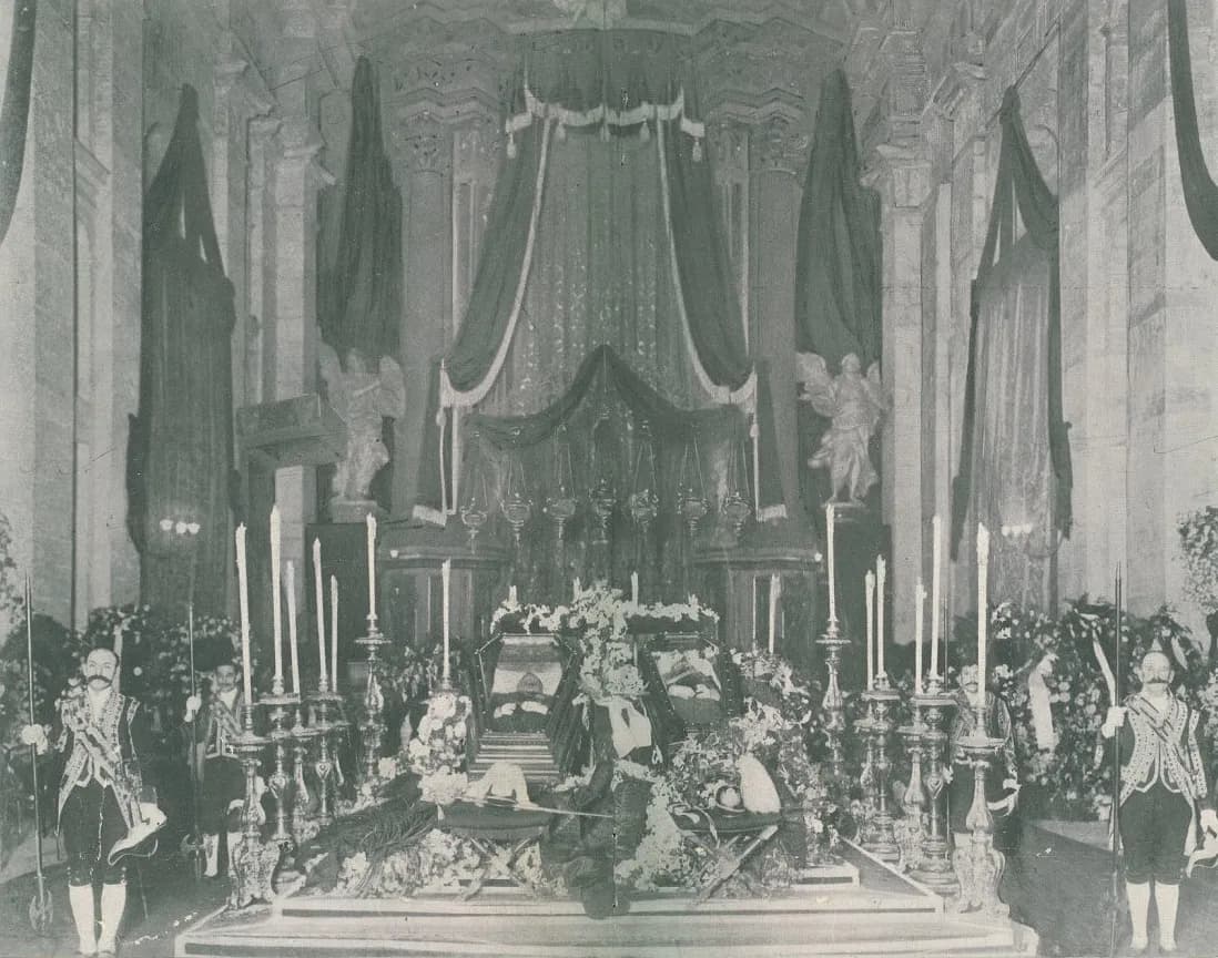 Two coffins heaped with flowers lie on a draped platform in a lofty church interior, surrounded by tall candlesticks. Guards in ceremonial uniform holding halberds stand at each side, and a canopy of dark hangings rises behind.