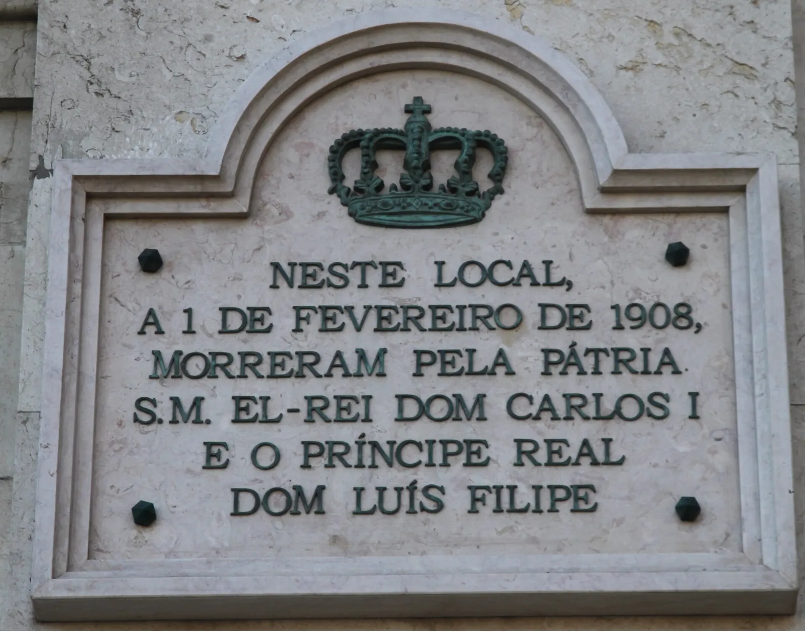 A pale stone plaque with an arched top, set into a wall, with a green bronze royal crown above raised capital letters in Portuguese.