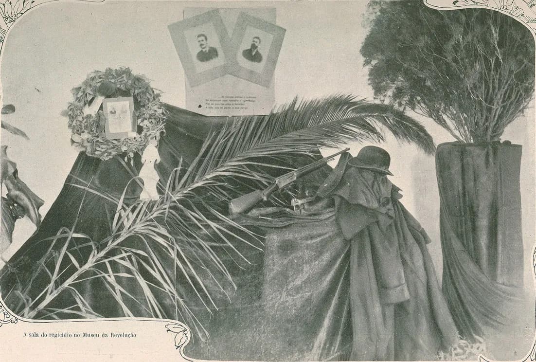 A display draped in dark cloth: a rifle and a pistol lie on a table beside a long cloak and a bowler hat, under a palm frond. Above them hang two framed portraits, one of a moustached man and one of a bearded man, and a laurel wreath circles a third picture.