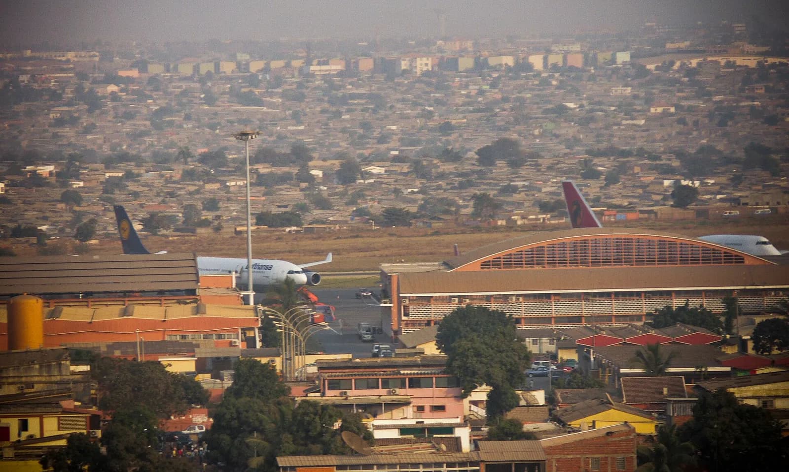 Aerial view of an airport terminal with a Lufthansa airliner and an Angola Airlines jumbo jet on the apron, and a dense spread of low houses beyond.