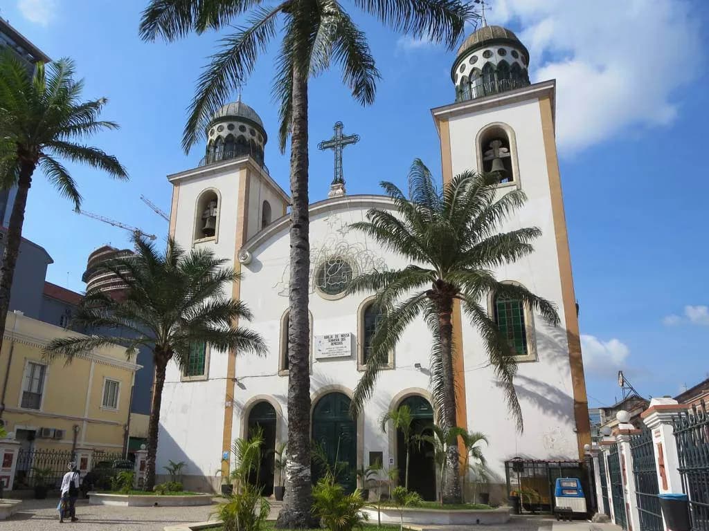 White church facade with two bell towers topped by small domes, a cross between them and palm trees in front, under a blue sky.