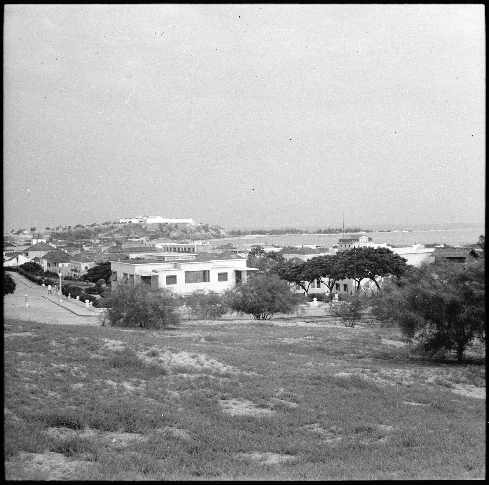 Black-and-white view over scrubby open ground, low white houses and trees towards Luanda's bay, with a white fortress on a hill in the distance.