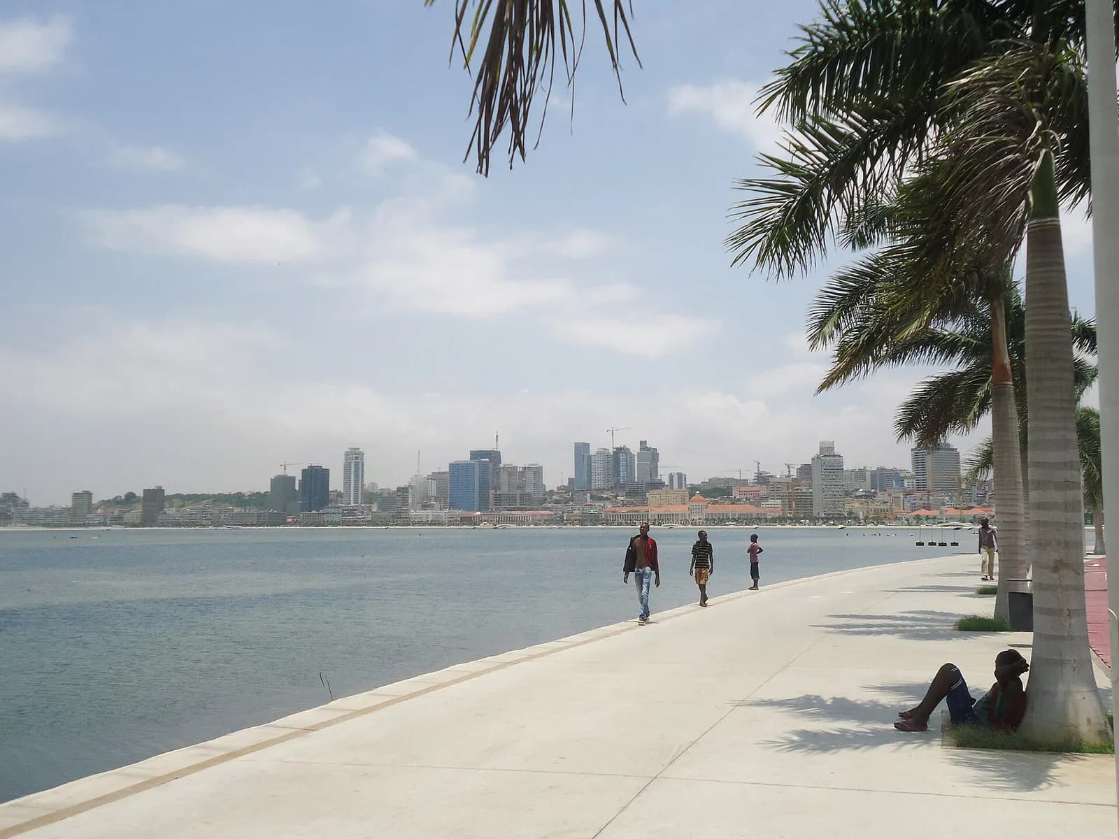 A wide paved promenade with palm trees runs along Luanda's bay, with a few walkers and the city's high-rise skyline across the water.