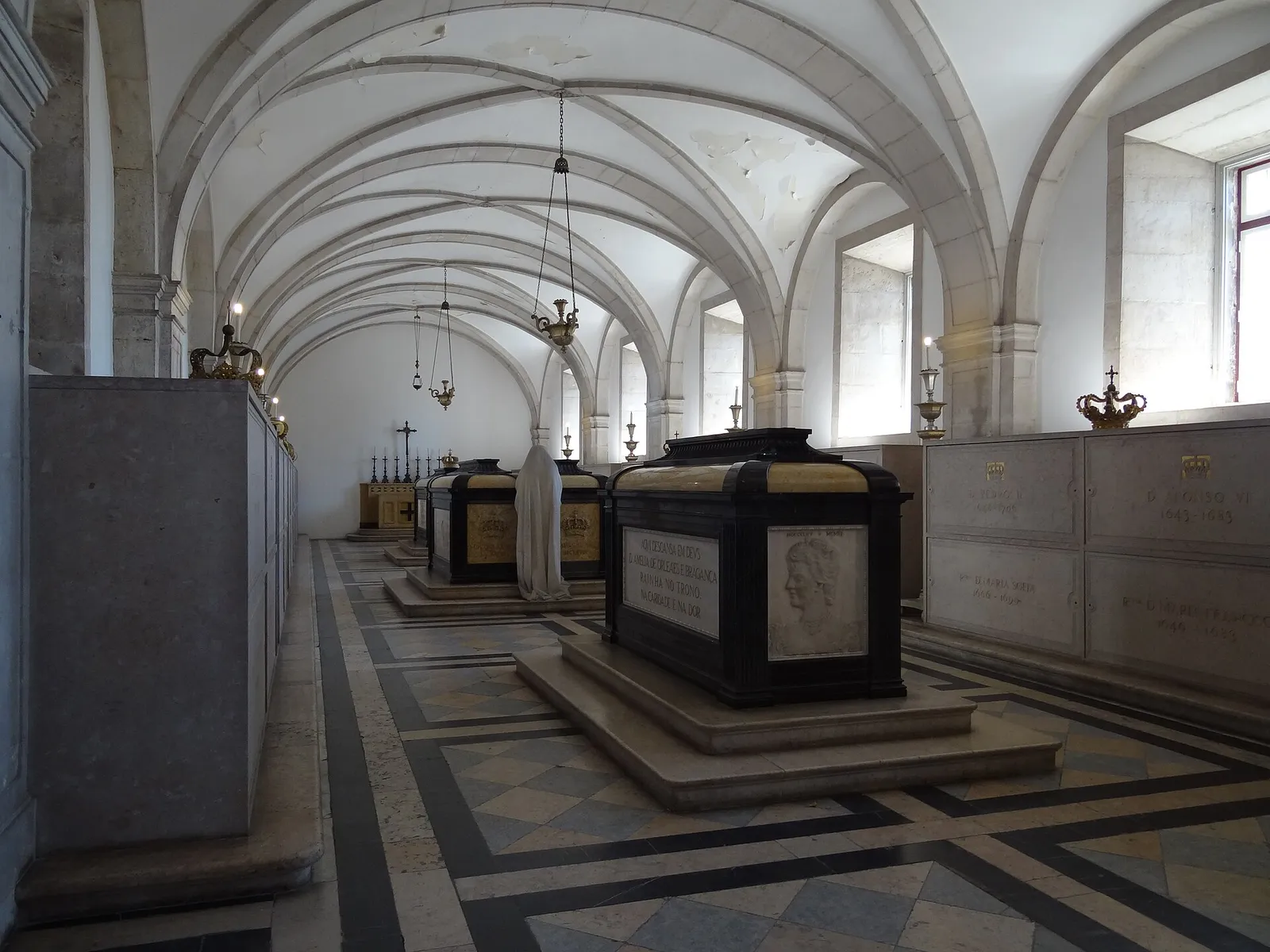 A long white vaulted hall with a patterned stone floor. Dark marble tombs with gilt crowns stand in the centre, and the side walls are lined with marble-faced burial niches, two of them inscribed D. Pedro II and D. Afonso VI.