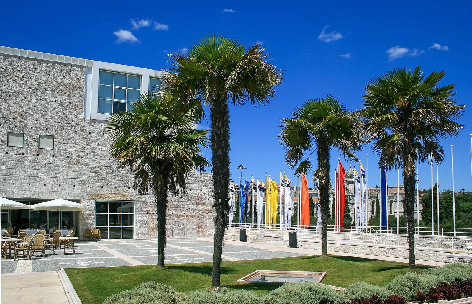 A pale stone building with small square openings in its walls stands beside a lawn with four palm trees and café tables. A row of tall coloured banners flies on flagpoles along a terrace under a deep blue sky.