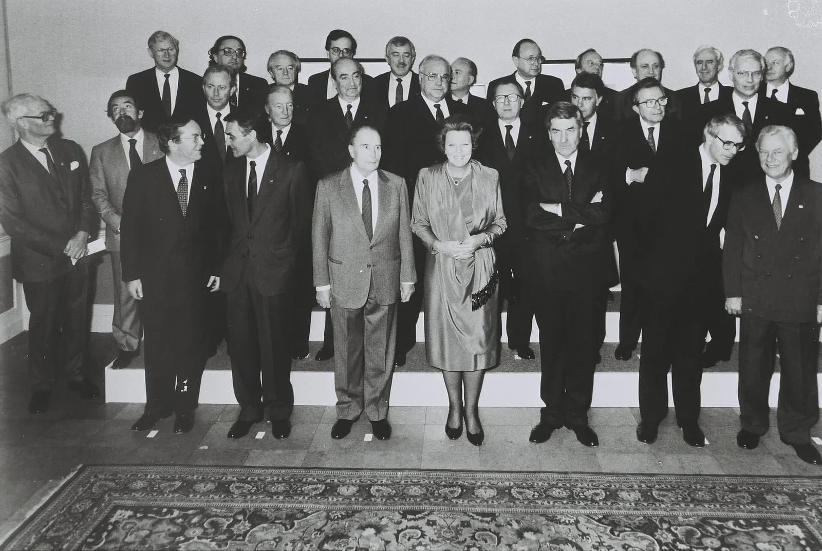 Black-and-white group photograph of more than twenty-five men in dark suits standing in three rows on low steps, with a woman in a light satin dress at the centre of the front row and a patterned carpet in the foreground.