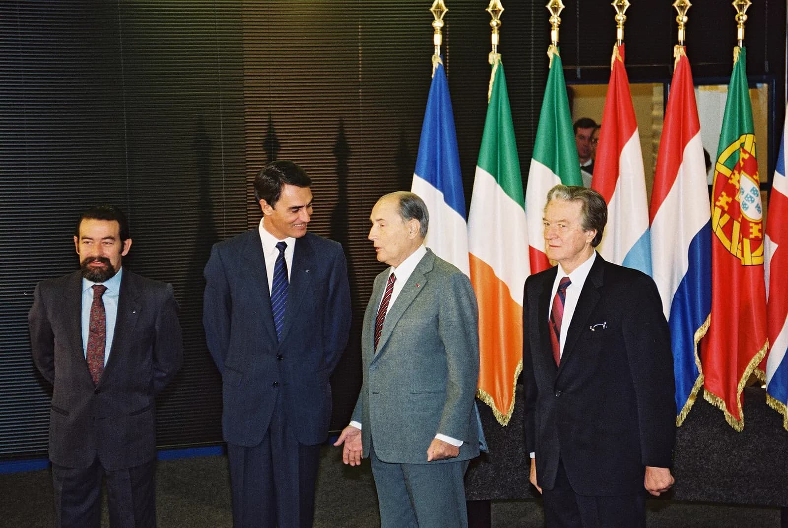 Four men in suits stand in front of a row of national flags, including the flags of France, Ireland, Italy, Luxembourg, the Netherlands, Portugal and the United Kingdom. A bearded man stands on the left, a tall dark-haired man smiles beside him, and an older man in a grey suit gestures as he talks.