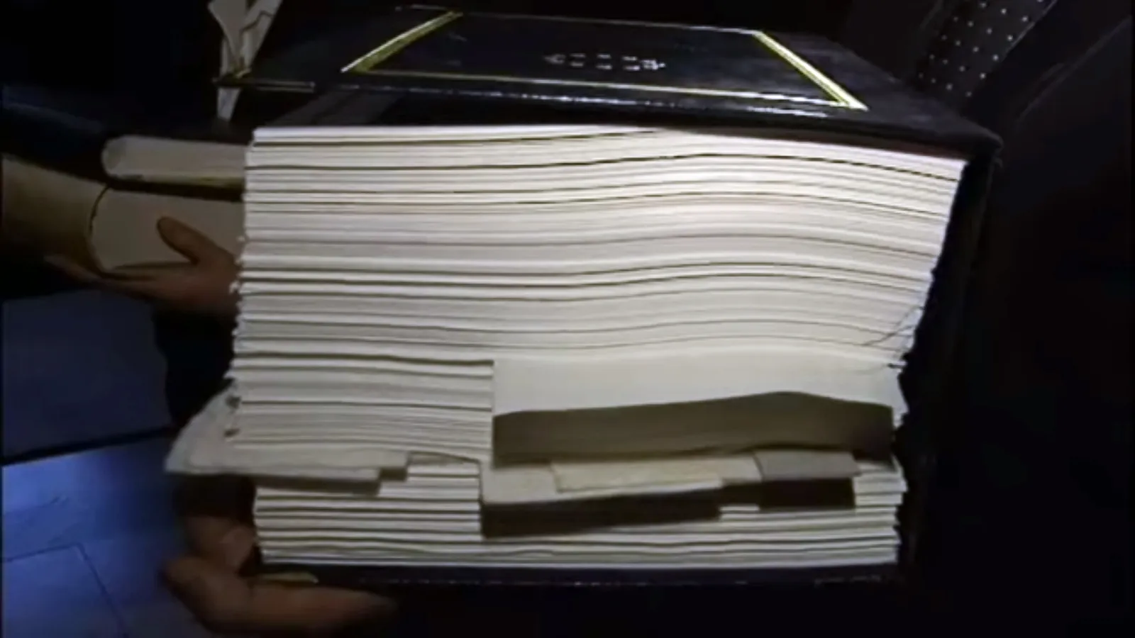 Hands hold open a very thick bound volume with a dark leather cover edged in gold, its hundreds of white pages and folded inserts seen side-on.