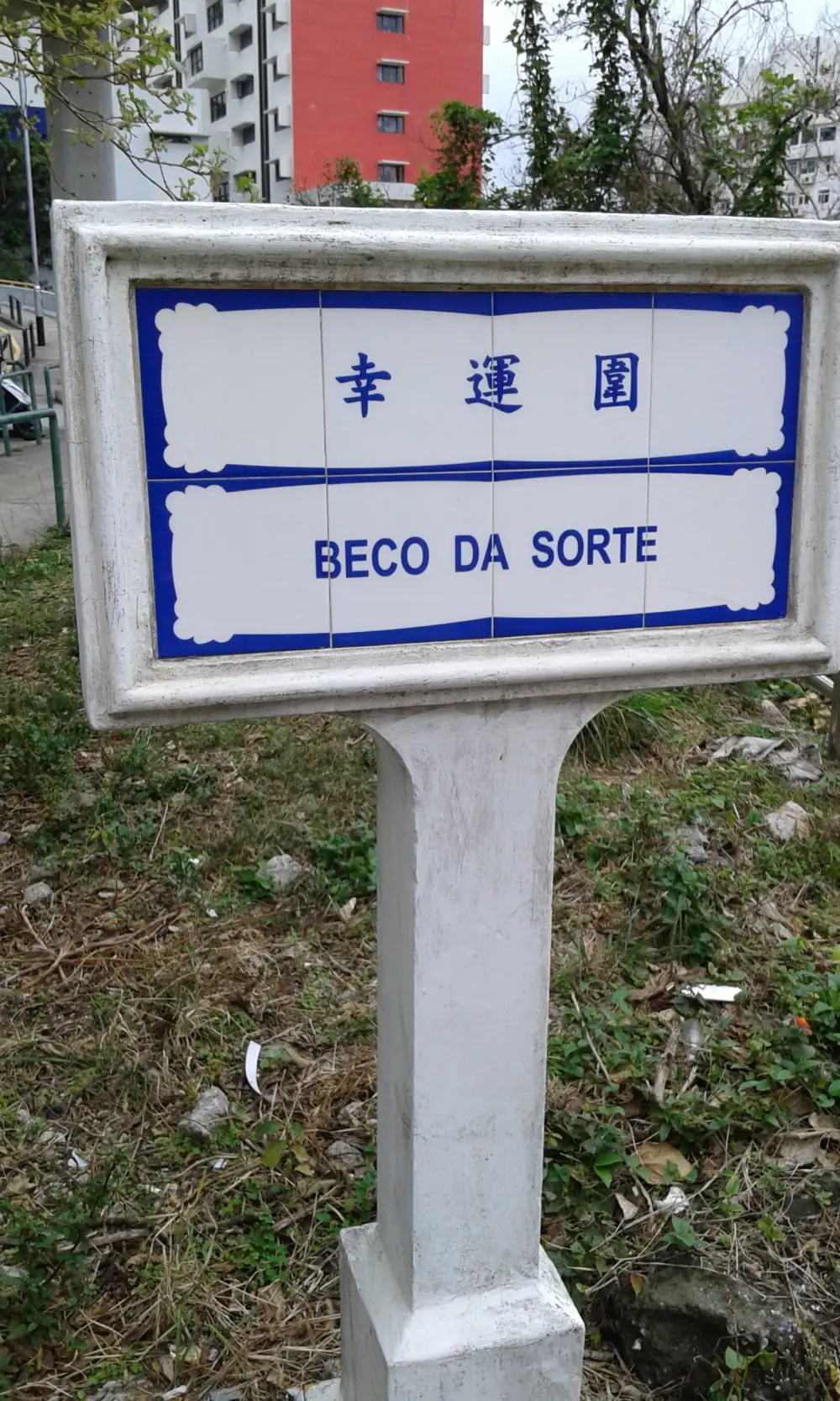 A white-framed tiled street sign on a post, with blue Chinese characters on the top row and the words Beco da Sorte in blue capital letters below, both inside a blue scalloped border. Trees and apartment blocks are in the background.
