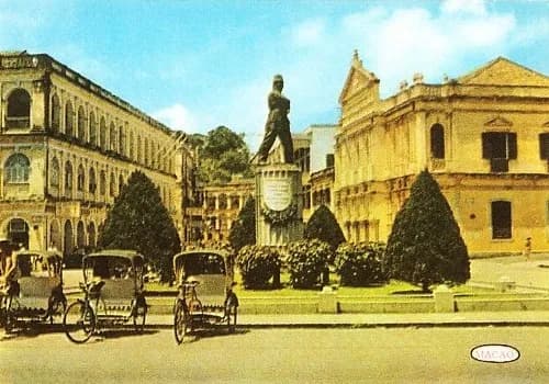 A coloured postcard of a bronze statue of a soldier on a tall pedestal in a square, flanked by trimmed conical trees and low hedges. Pale arcaded colonial buildings stand on both sides, and three empty cycle rickshaws wait in the foreground.