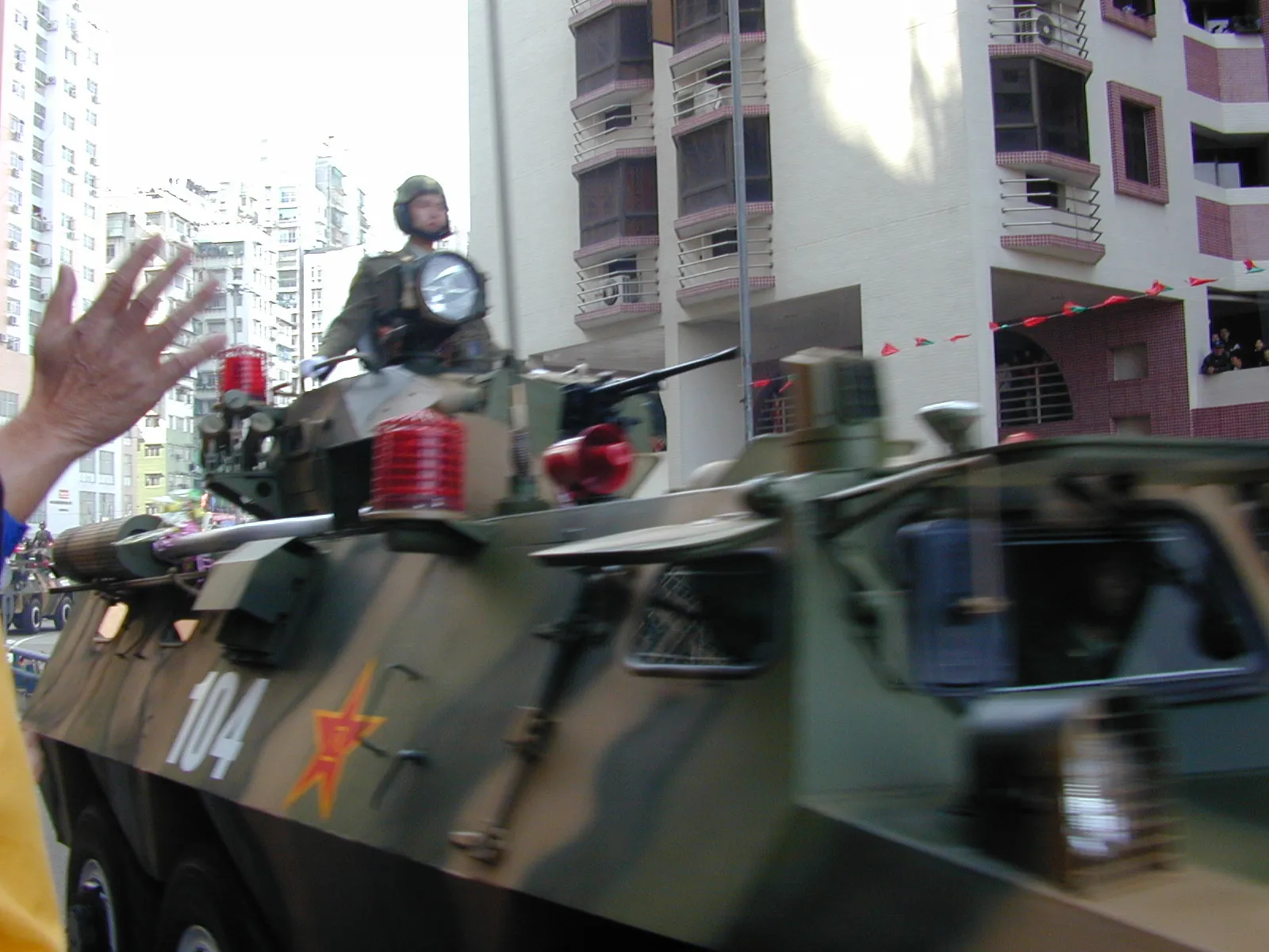 A camouflaged armoured personnel carrier marked with a red star and the number 104 drives past apartment blocks hung with small flags. A helmeted soldier in white gloves stands in the hatch, and a spectator's raised hand waves in the foreground.