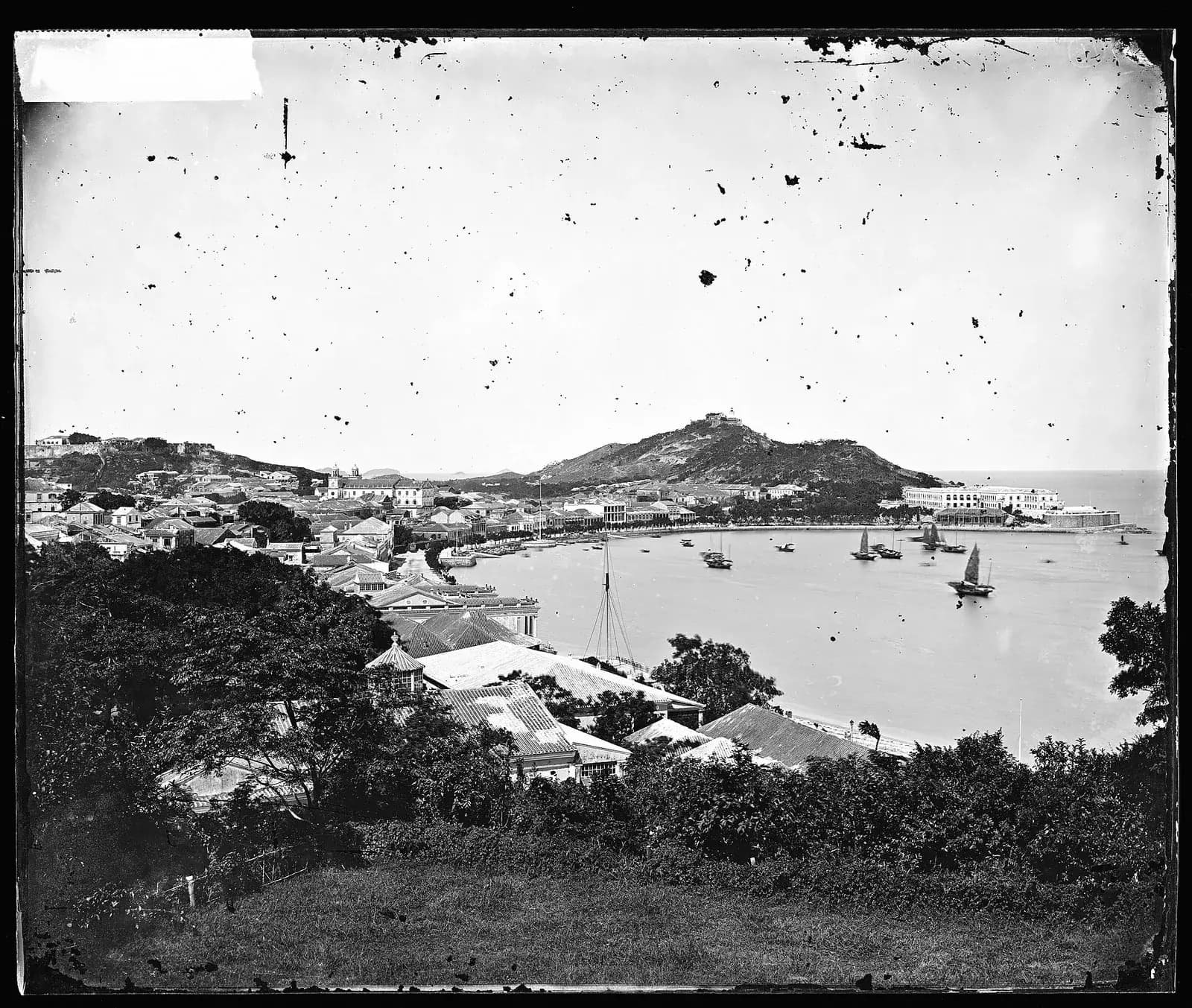 A black and white view from a wooded hillside over a curving bay lined with low houses and a waterfront promenade. Sailing junks and small boats float on the bay; a steep hill with a building on its summit rises behind the town, and a long white building stands at the far end of the shore.