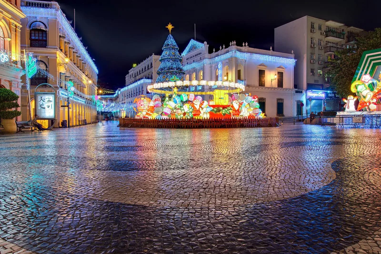 A wide square paved with black and white stones laid in a wave pattern, wet and shining at night. Arcaded colonial buildings strung with blue and white lights line the left side; at the back stands a white building with a pediment, and in front of it a tall lit Christmas tree and a fenced display of glowing lanterns.