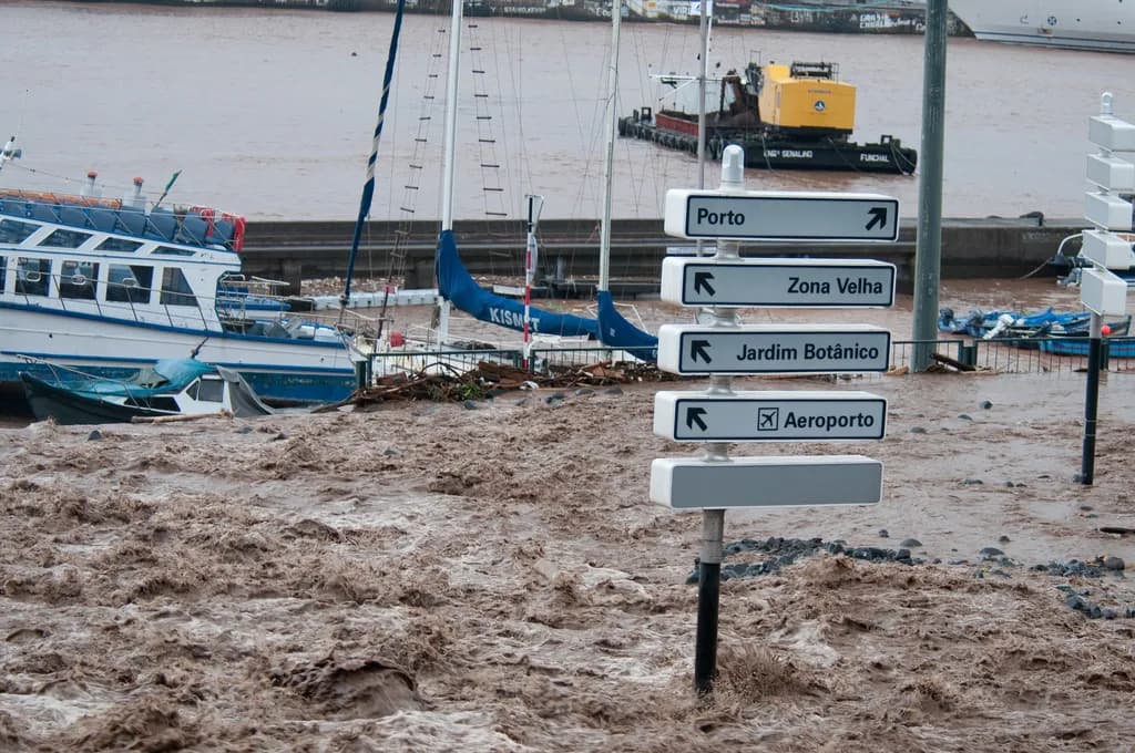 A churning sheet of brown floodwater covers a seafront road. A white signpost pointing to Porto, Zona Velha, Jardim Botânico and Aeroporto stands in the current, and behind it moored sailing boats, a tourist boat and a yellow dredger sit in muddy harbour water.