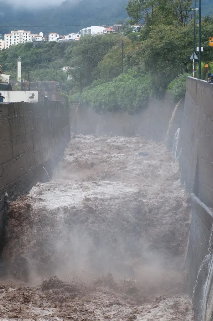A torrent of brown water and spray fills a narrow channel between high walls, with trees and white houses on the hillside above.