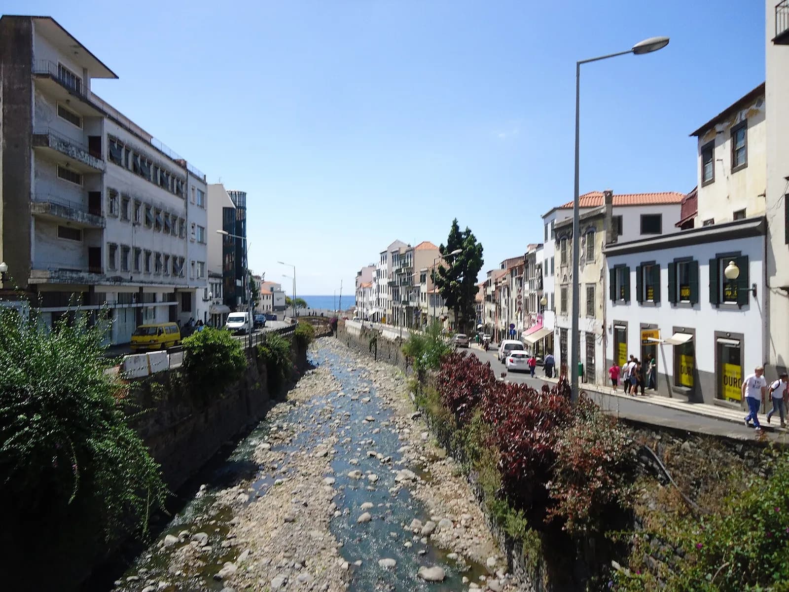 A shallow stream runs over grey stones between high stone walls in the middle of a city street. Shrubs grow along the tops of the walls, and pavements, parked cars and white buildings line both sides, with the sea at the far end.