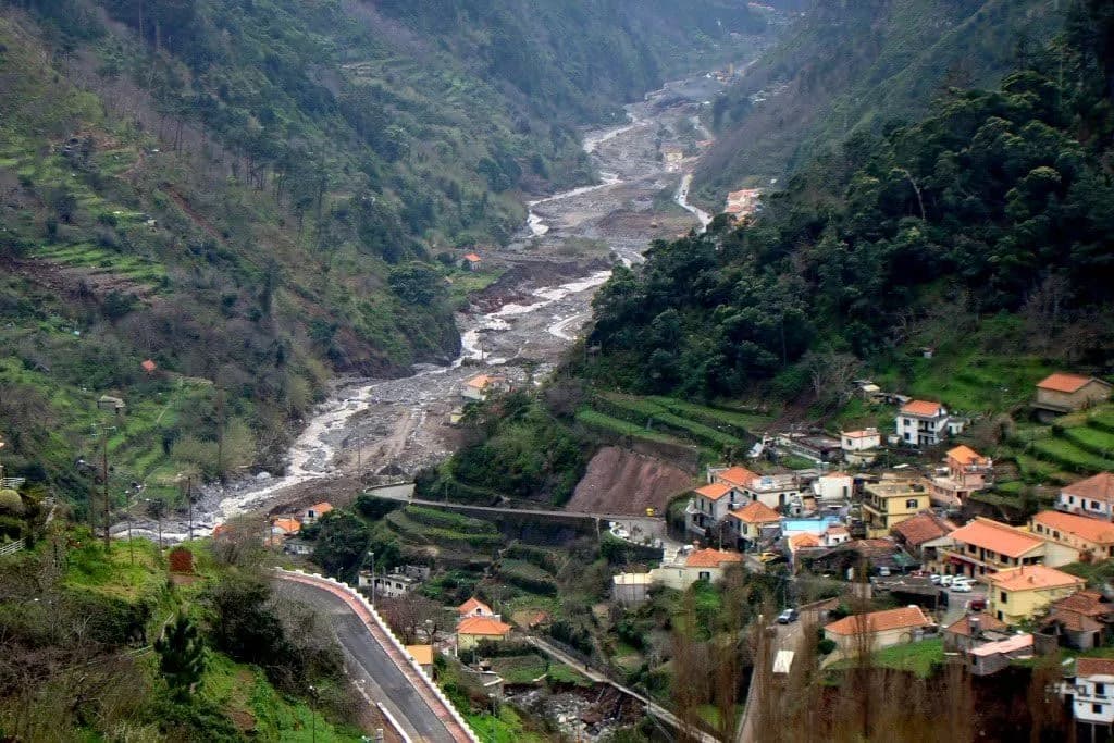 A steep green valley with terraced slopes. Its floor is a wide band of grey rocks and rubble where the stream has torn out its banks, and a cluster of orange-roofed houses and a winding road sit on the hillside to the right.