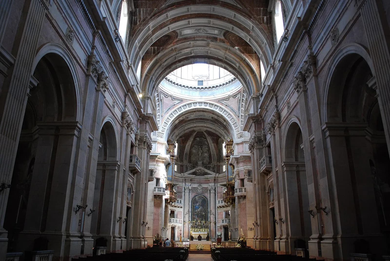 Inside a tall stone church: a long nave of grey and pink marble, with arched side chapels and small balconies, leads under a coffered vault to a domed crossing and a high altar with a large painting.