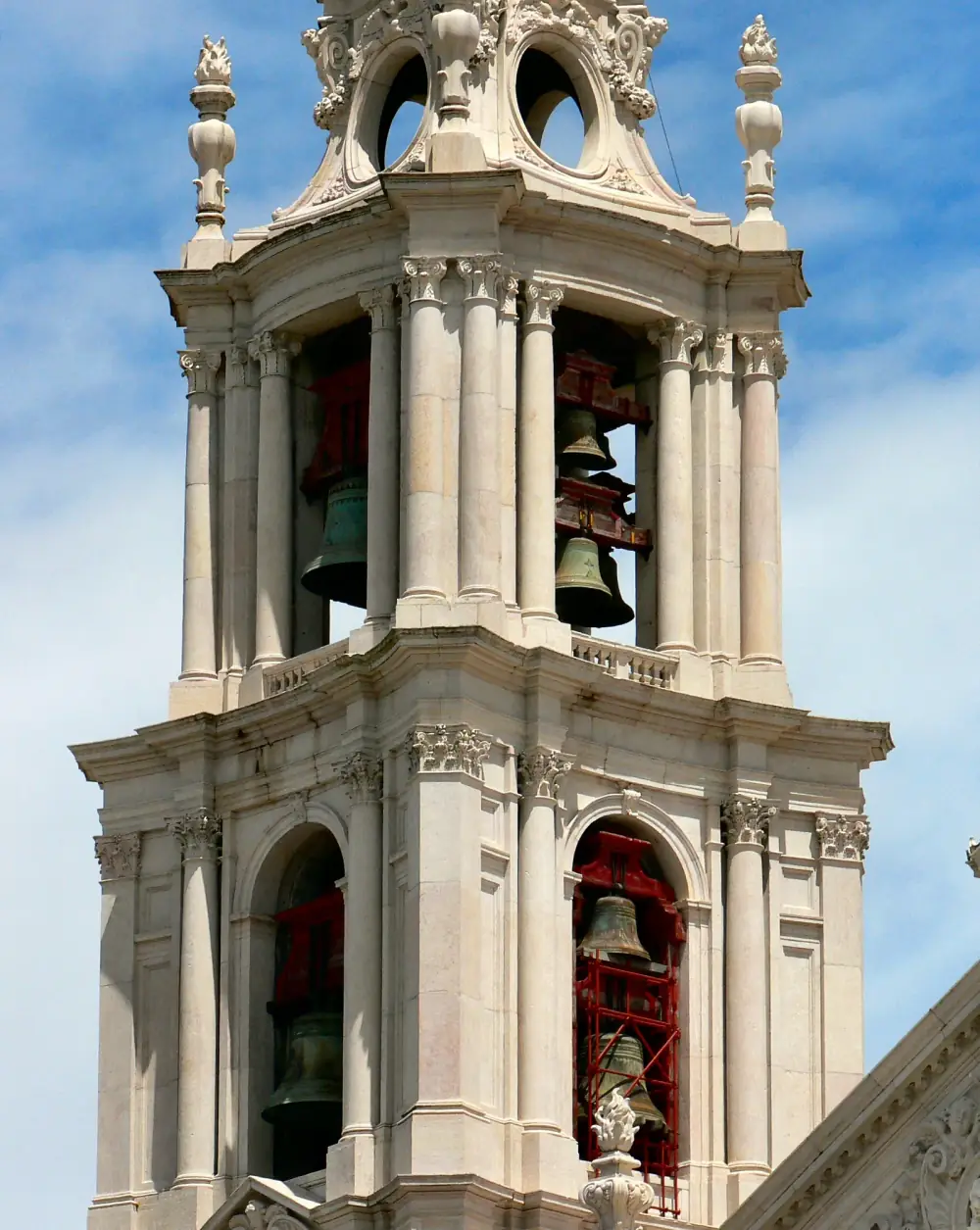 The top of a pale limestone bell tower against a blue sky, with Corinthian columns and stone finials. Large bronze bells hang in red-painted frames behind its arched openings.