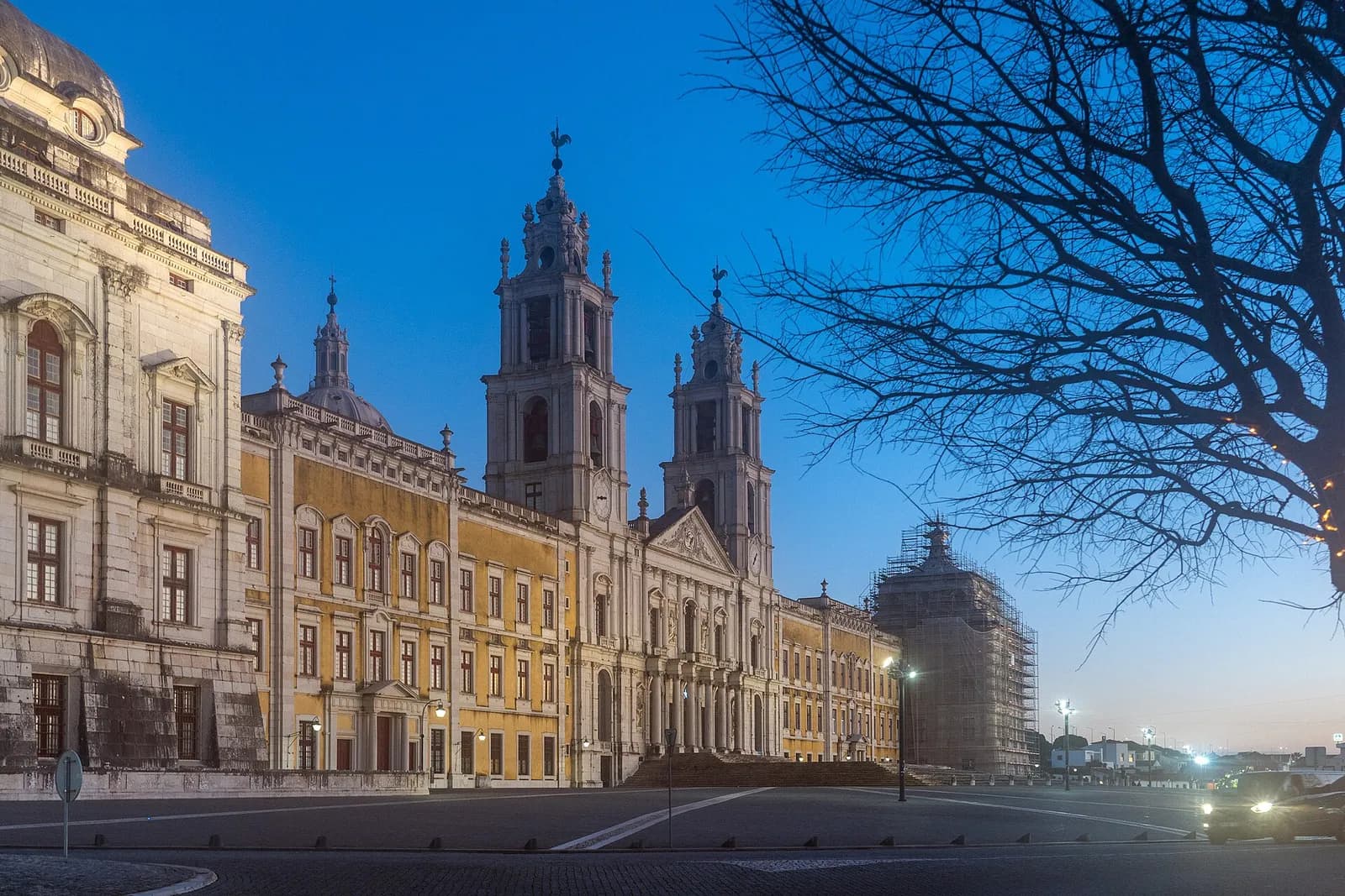 The long palace façade at dusk under a deep blue sky: a pale stone corner pavilion on the left, ochre-painted wings, and in the middle a church front between two tall white bell towers. A pavilion on the far right is wrapped in scaffolding, and bare branches cross the right of the picture.
