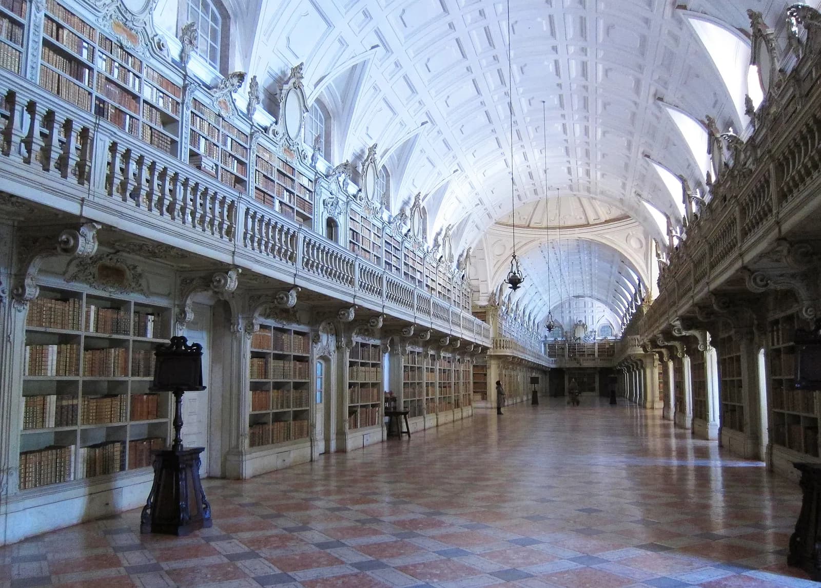 A long hall with a white coffered barrel vault and a chequered pink and grey marble floor, lined on both sides with wooden bookcases full of leather-bound books, with an upper gallery behind a balustrade.