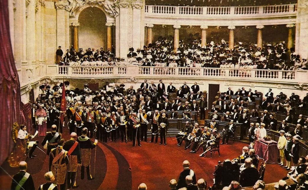 A coloured photograph of a semicircular parliamentary hall. Men in dark uniforms, sashes and court dress stand on a red floor in front of tiered wooden benches, and women in pale hats fill the galleries above them.
