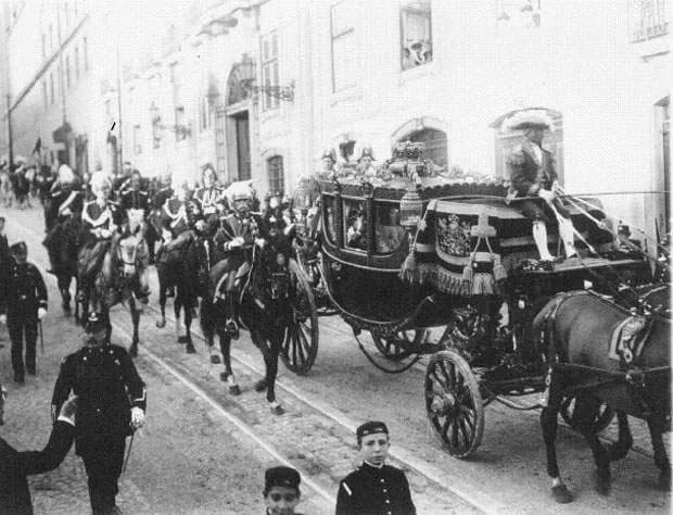 A gilded state coach drawn by horses moves along a cobbled street between tall buildings, escorted by mounted soldiers in plumed helmets. A coachman in a plumed hat sits up front; a policeman and two boys in caps stand in the foreground.