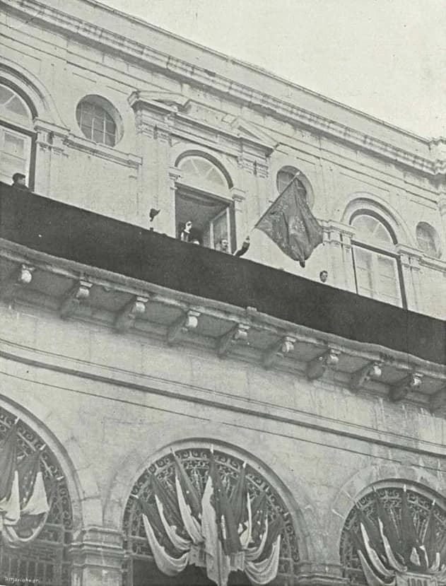 The upper floor of a stone building with arched windows. On a long balcony draped in dark cloth, a few men stand beside a large flag on a staff; below, the arches are hung with festoons of flags.