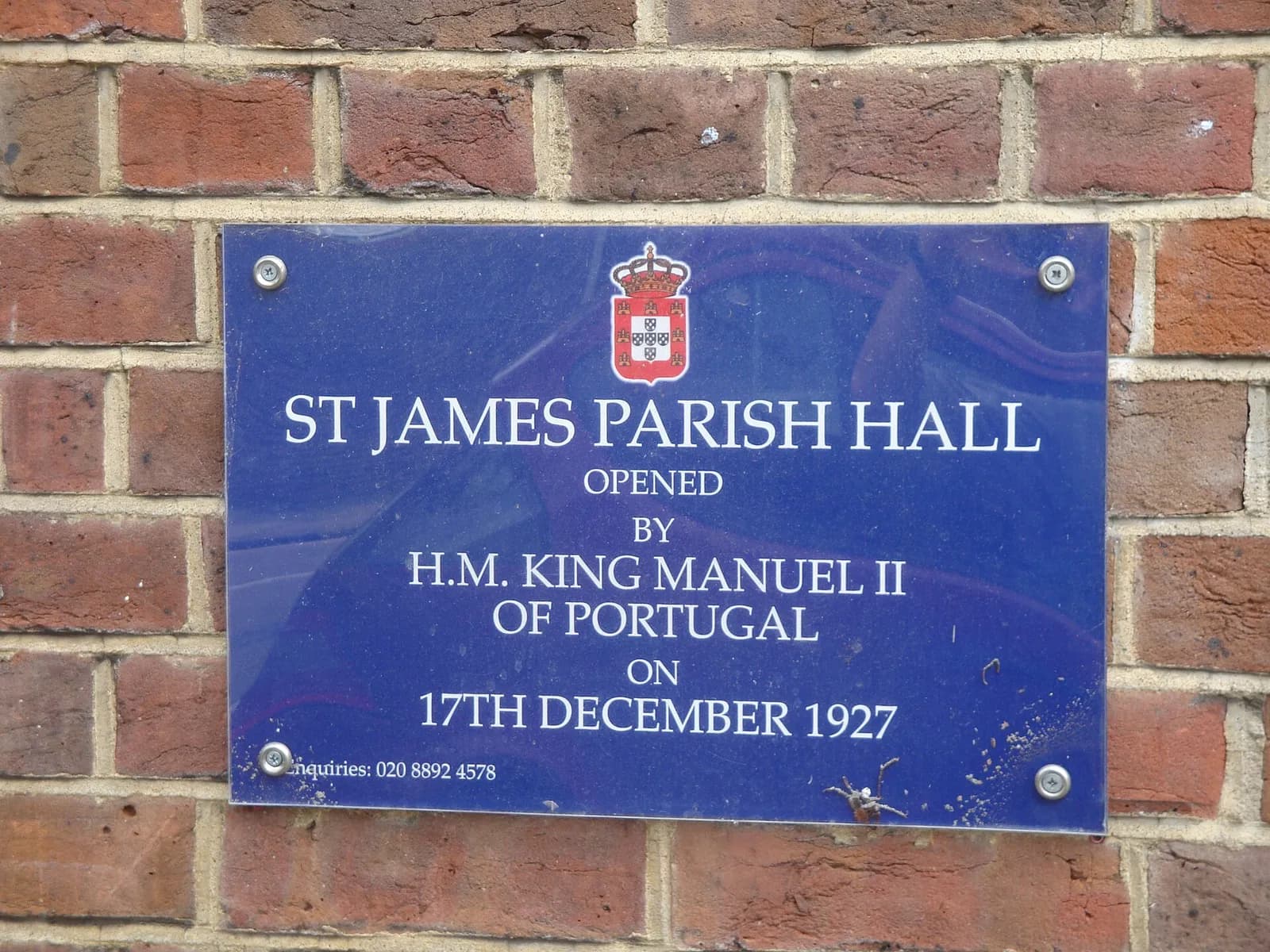 A blue plaque on a red brick wall, topped by a small crowned Portuguese coat of arms, reading: St James Parish Hall, opened by H.M. King Manuel II of Portugal on 17th December 1927.