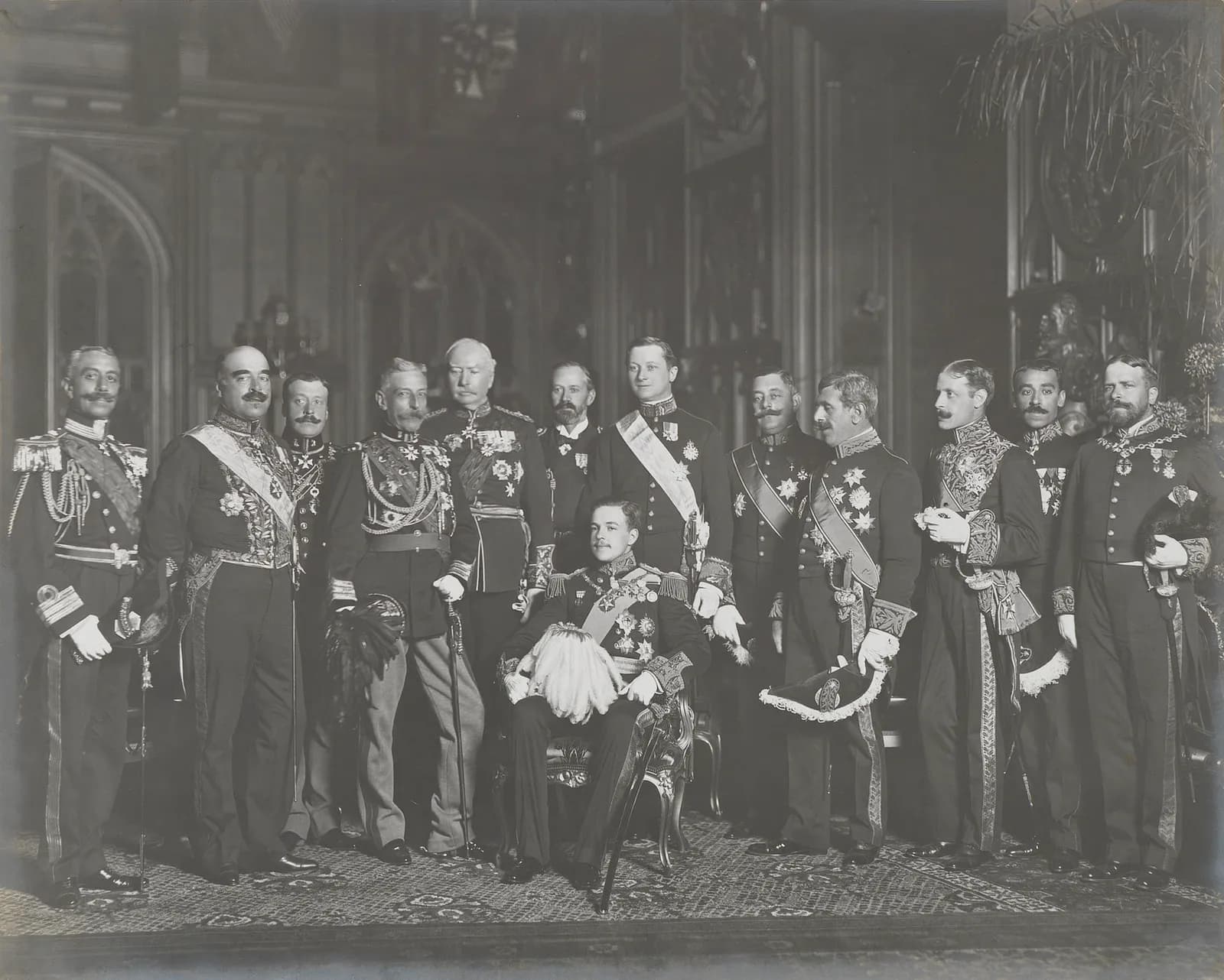 A formal group photograph of thirteen men in court dress and uniforms with sashes, stars and swords, standing in a panelled hall. A young man in uniform, holding a plumed hat, sits in a chair in the front centre.