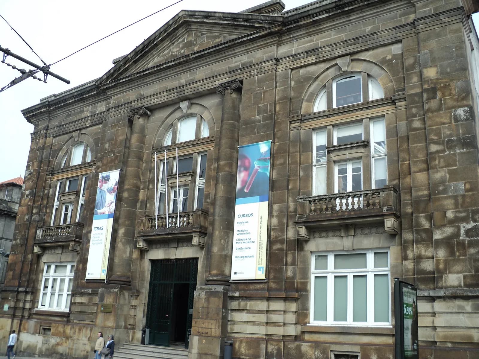 The weathered stone facade of an older building in Porto with a triangular pediment, two columns, arched windows and small balconies. Two tall banners hang on the front, one with the name of ICBAS and one listing its courses. A few people walk past on the pavement.