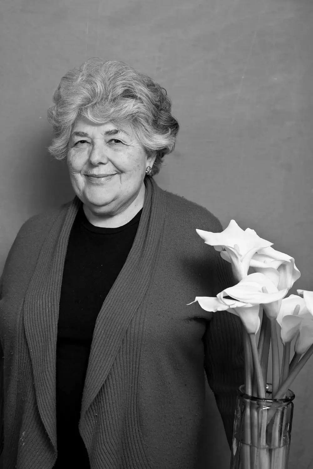 Black and white portrait of an older woman with short wavy grey hair, smiling, wearing a dark top and a long knitted cardigan, standing beside a glass vase of white calla lilies.