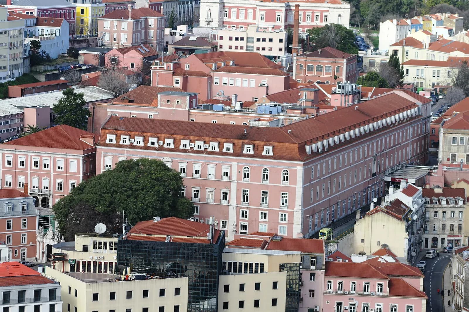 View over central Lisbon rooftops towards a long pink building with a red tiled roof and rows of dormer windows, surrounded by other pastel buildings and trees.