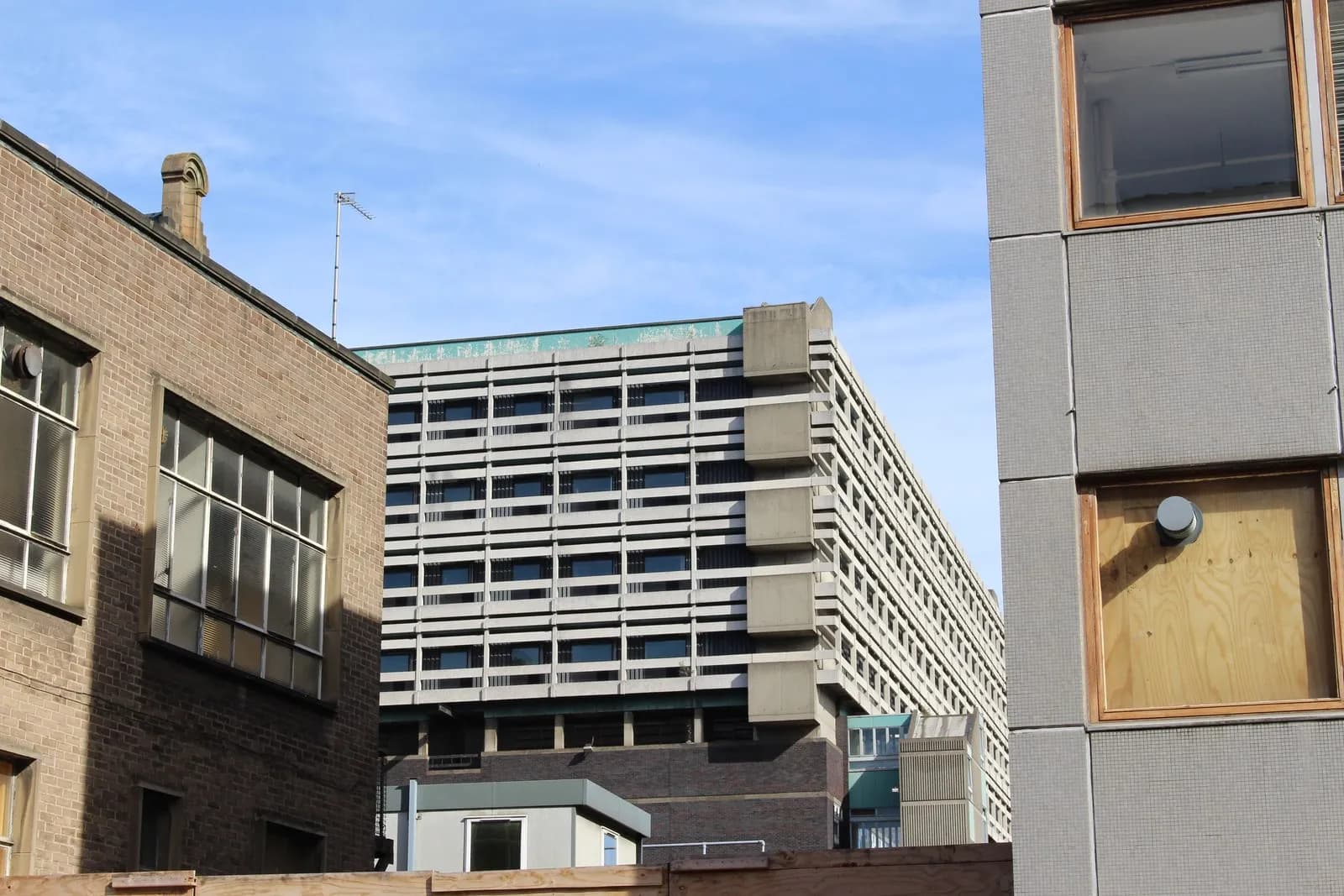 A tall concrete hospital block with horizontal bands of windows rises behind older brick and stone buildings under a blue sky.